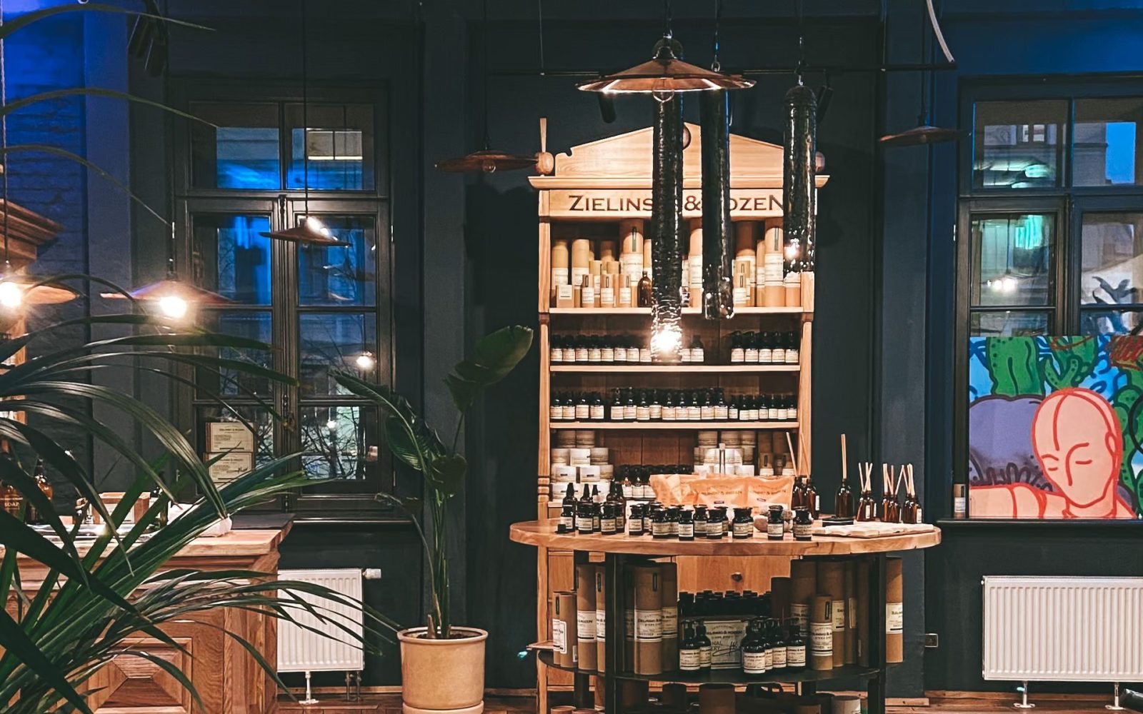 Winkelinterieur met een houten kast en een ronde tafel vol verzorgingsproducten, tussen planten en hanglampen